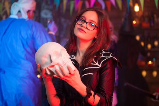 Pretty little girl dressed up like a witch holding a human skull at halloween party. Creepy man dressed up like a doctor in the background.