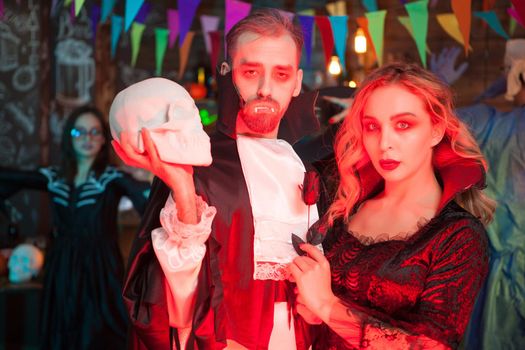 Portrait of terrifying zombie couple at halloween celebration. Man dressed up like dracula holding a human skull.