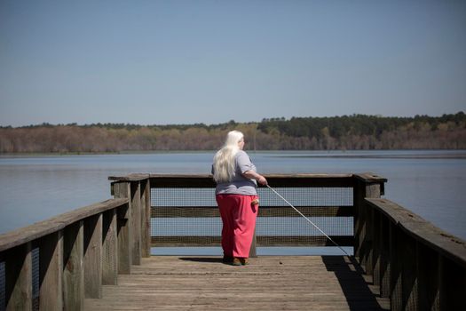 Blind woman with white hair standing at the edge of a pier near water and holding a cane