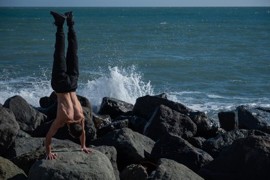 Shirtless man doing handstand on rocks by the sea