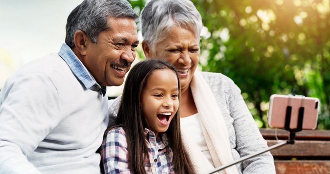 Capturing all the best moments. a cheerful little girl sitting down on a bench and taking a selfie with with her grandparents at the park