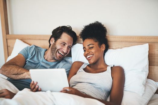 Happy, relaxed and carefree couple reading social media news on digital tablet and laughing in bed. Interracial husband and wife waking up together and browsing internet, sharing a funny online joke.