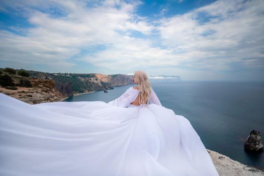 Blonde with long hair on a sunny seashore in a white flowing dress, rear view, silk fabric waving in the wind. Against the backdrop of the blue sky and mountains on the seashore