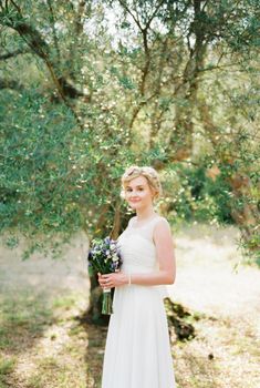 Bride with a bouquet of flowers stands under an olive tree. High quality photo