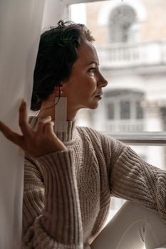 Free time for rest. Profile of a beautiful woman sitting on a white window sill at home, holding a cotton plant in her hands. She is wearing a sweater, leggings and white boots