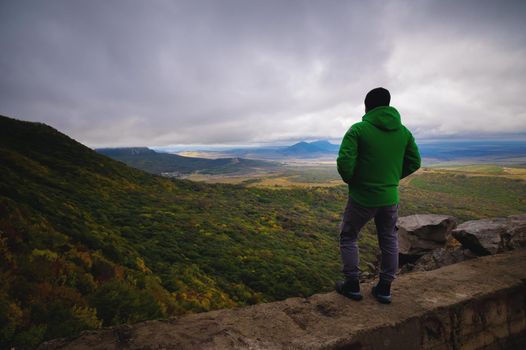 a man traveling on foot, standing in a bright jacket on top of one of the mountains, looking into the distance at the mountains and the cloudy sky.