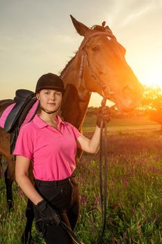 Beautiful smiling girl jockey stand next to her brown horse wearing special uniform on a sky and green field background on a sunset. Equitation sport competition and activity.