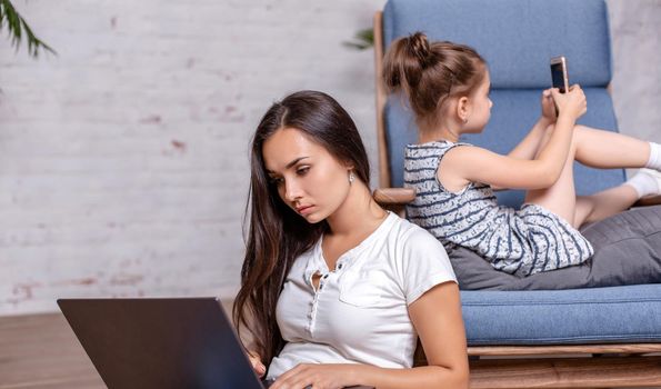 Mother working with laptop, sitting on the floor and cute little girl laying on the armchair and playing games on the smartphone