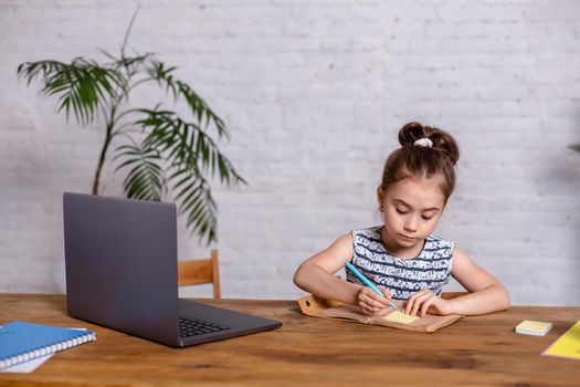 Cute little girl is sitting at table with her laptop and notebook. The concept of new programs for teaching children
