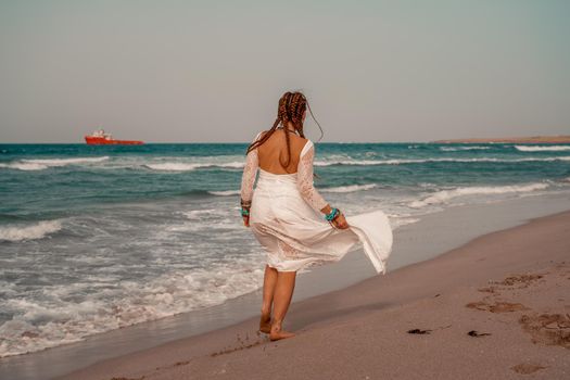 Model in boho style in a white long dress and silver jewelry on the beach. Her hair is braided, and there are many bracelets on her arms