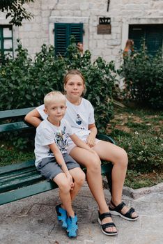 A little boy with long hair and a girl are sitting on a park bench. Children talk while sitting in a pack on a bench. Boy and girl spend their free time outdoors. Portrait of a girl and a boy.