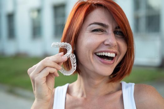 Red-haired Caucasian woman holding transparent mouthguards for bite correction outdoors. A girl with a beautiful snow-white smile uses silicone braces.