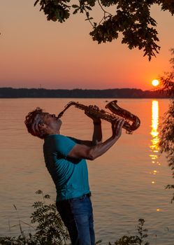 An adult man plays the saxophone at sunset by the river in the evening.