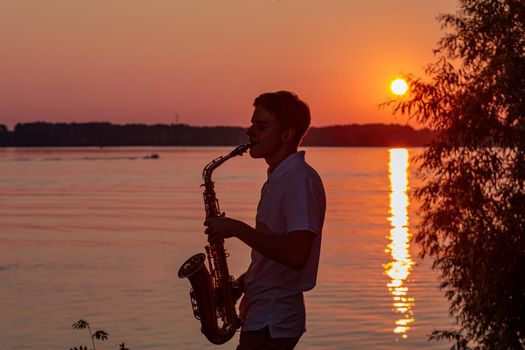 A young guy plays the saxophone in the evening at sunset.