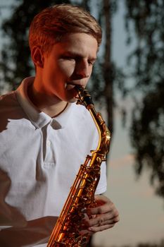 A young guy plays the saxophone in the evening at sunset.