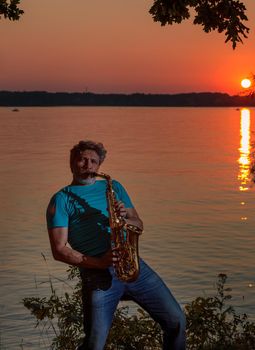 An adult man plays the saxophone at sunset by the river in the evening.