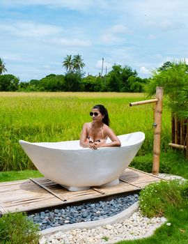 Asian women in a white bathtub outside looking out over a green rice field, men and women in bath tub outside on vacation at a homestay in Thailand with green rice paddy field