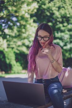 Young girl with pink hair is studying in the spring park, sitting on the wooden bench and browsing on her laptop