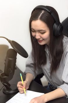 Young millennial woman with professional microphone and headphones taking notes and recording podcast at studio, technology and media concept, vertical image, selective focus