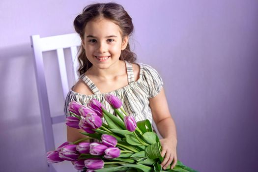 Beautiful little girl with a bouquet of tulips, sits on a white chair, against a purple wall, smiles, looks at the camera. Copy space