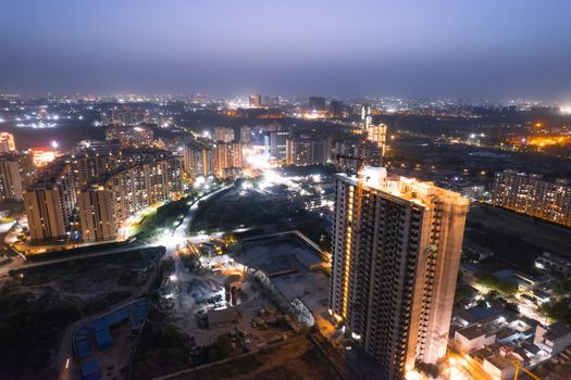 aerial drone dusk twilight shot showing orange lights of streets, homes and markets surrounding a skyscraper with the city scape stretching into the distance in gurgaon haryana delhi drone shot