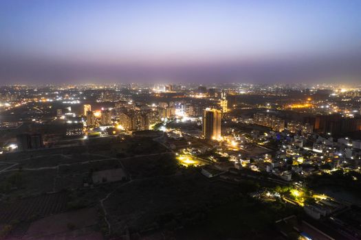 aerial drone dusk twilight shot showing orange lights of streets, homes and markets surrounding a skyscraper with the city scape stretching into the distance in gurgaon haryana delhi drone shot