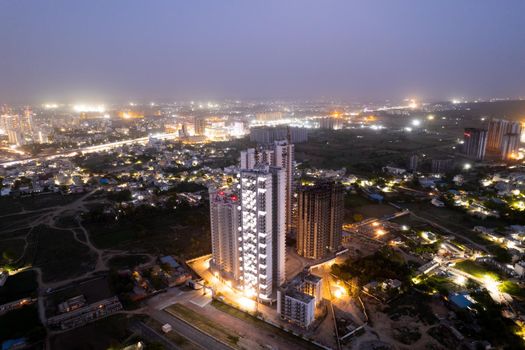 aerial drone dusk twilight shot showing orange lights of streets, homes and markets surrounding a skyscraper with the city scape stretching into the distance in gurgaon haryana delhi drone shot