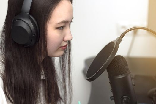 Young millennial woman in front of professional microphone with headphones, closeup, recording of podcast at studio, technology and media audio concept