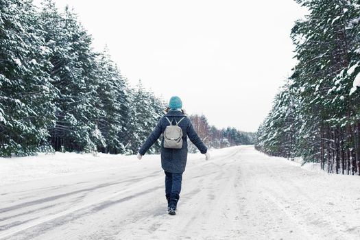 a girl in a gray coat and a blue knitted hat, with a gray backpack on her back, walks along the side of a winter road, next to a beautiful winter forest on a cloudy snowy day. Back view