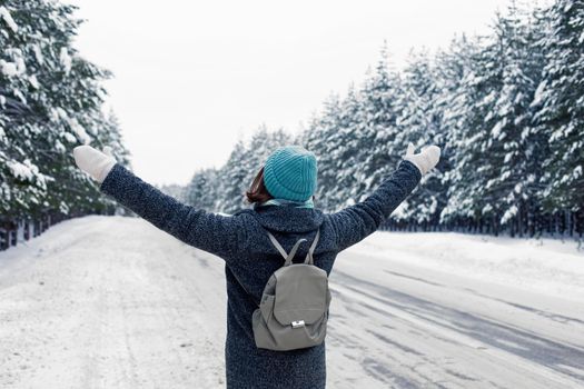 a girl in a gray coat and a blue knitted hat, with a gray backpack behind her back, stands on the side of the road, in a beautiful winter snowy forest on a cloudy snowy day, raising her hands. Back view