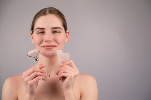 Caucasian woman holding pink roller massager and gouache scraper on white background. Copy space