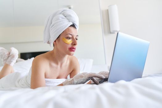 After a shower, a girl wrapped in a towel works on a laptop and uses cosmetic patches for the skin under the eyes, lips and gloves to moisturize her hands and feet. Cosmetic trends for body care at home.
