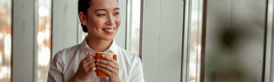 Smiling asian business woman drinking coffee standing near window at office. High quality photo