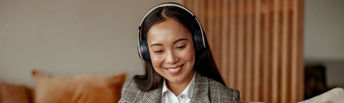 Asian business woman listening music on headphones sitting at laptop with coffee in cozy office