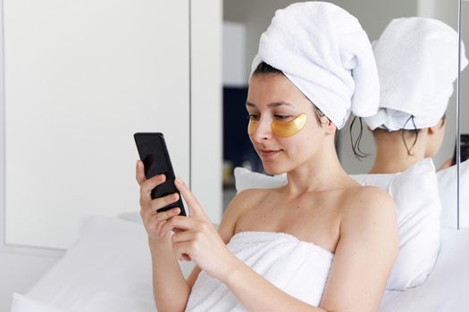 After a shower, the girl is wrapped in a towel and wears cosmetic patches for the skin under the eyes. Looks at the cell phone. Cosmetic procedures at home. Getting ready for a date.
