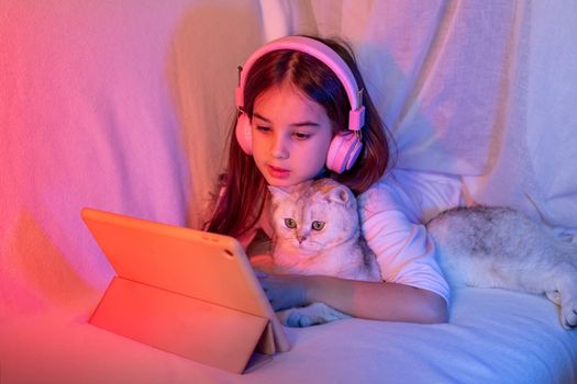 A beautiful little girl in pink headphones, with loose dark hair, lies on the bed, with a white adorable cat, in a neon pink-blue light, looks into a digital tablet
