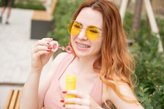 Young red-haired woman blowing soap bubbles outdoors. Girl in yellow sunglasses and braces