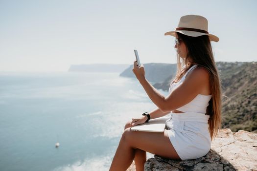 Successful business woman in yellow hat working on laptop by the sea. Pretty lady typing on computer at summer day outdoors. Freelance, travel and holidays concept.