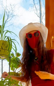 A girl in a red dress, a white hat, funny sunglasses and a red medical mask with plants against the sky in early spring