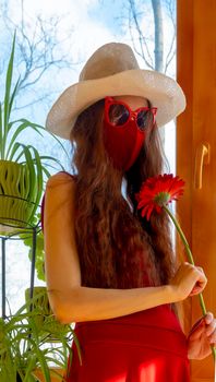 A girl in a red dress, a white hat, funny sunglasses and a red medical mask is hold red flower with plants against the sky in early sprin