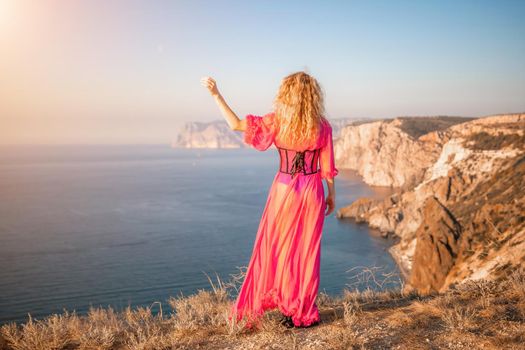 Side view a Young beautiful sensual woman in a red long dress posing on a volcanic rock high above the sea during sunset. Girl on the nature on blue sky background. Fashion photo