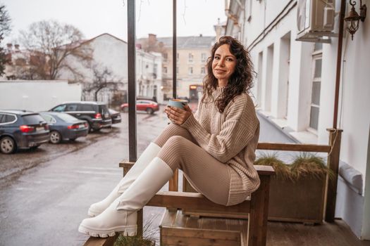 A middle-aged woman in a beige sweater with a blue mug in her hands is in a street cafe on the veranda.