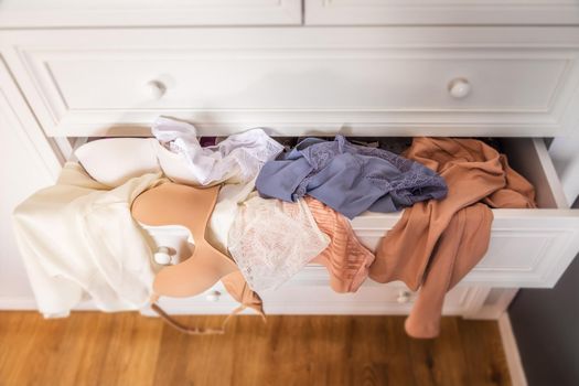 close-up drawers with clothes in white closet in the apartment are pulled out, top view Lingerie, lace panties, bra and t-shirt in a mess on shelf. Sun rays in the morning, a bouquet of flowers nearby