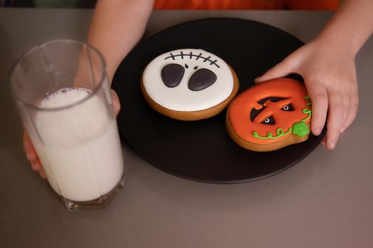 close-up, pumpkin gingerbread cookies on a plate, next to a glass of milk. Close-up, the girl's hand takes a cookie. Halloween holiday concept, home party, handmade pastries