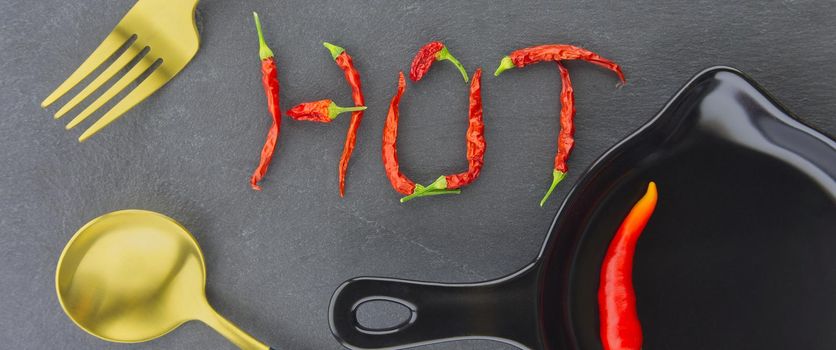 HOT spicy chili pepper still life with text formed from dried pods, gold eating utensils and a fresh chilli pepper in a skillet in a panorama banner over grey
