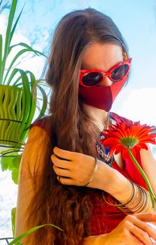 A girl in a red dress, red medical mask and funny sunglasses holds a red gerbera flower