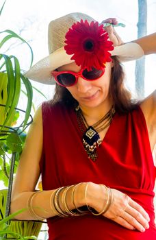 A girl in a red dress, white hat and funny sunglasses tries on a red gerbera flower as a hat decoration