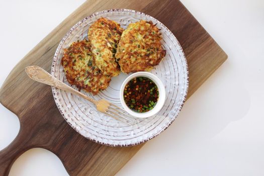 vegetarian zucchini fritters in a white plate and asian sauce with spices in a small white bowl on a wooden cutting board. White background. Vegetarian food concept