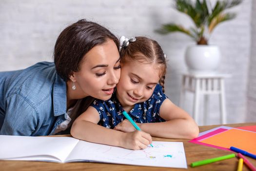Attractive young woman and her little cute daughter are sitting at the table and having fun while doing homework together. Mother helps daughter with her school classes.
