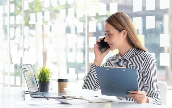 Portrait of young asian business woman talk on smartphone write notebook laptop in home office. Beautiful girl at desk computer take home order. Startup business asia woman online sme telemarketing..